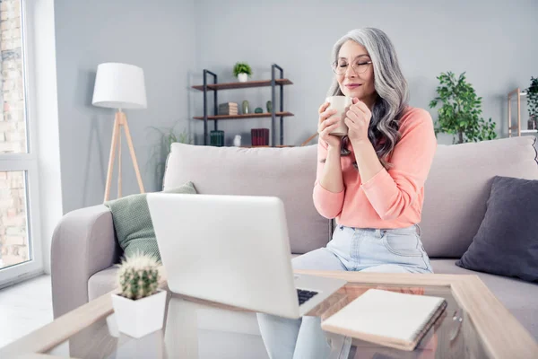 Portret van aantrekkelijke dromerige vrouw van middelbare leeftijd met behulp van laptop zittend op divan drinken cacao rustpauze thuis huis flat indoor — Stockfoto