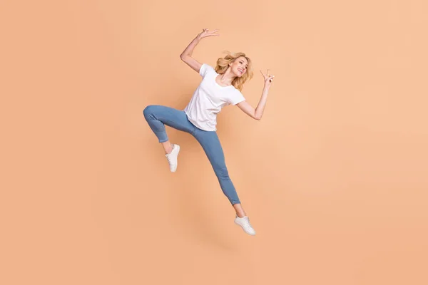 Foto van charmante glanzende dame dragen witte t-shirt glimlachen springen hoog tonen v-teken op zoek naar lege ruimte geïsoleerde beige kleur achtergrond — Stockfoto