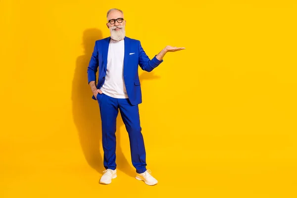 Full storlek foto av positiva grått skägg gammal man hålla tomt utrymme slitage glasögon blå jacka isolerad på gul bakgrund — Stockfoto