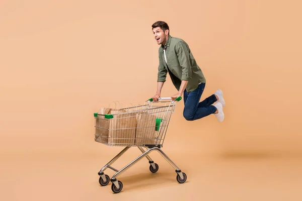 Photo of funny sweet young guy dressed green shirt smiling doing purchases riding shopping trolley isolated beige color background — 图库照片