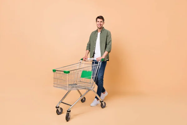 Full size photo of charming young happy man hold pushcart smile shopper isolated on beige color background — ストック写真