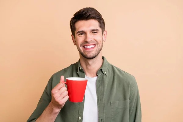 Фото привабливого молодого щасливого чоловіка тримає ручну чашку кави гарного настрою посмішка ізольована на бежевому кольоровому фоні — стокове фото