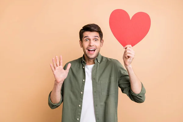 Foto de encantador bonito jovem cavalheiro desgaste cáqui camisa sorrindo segurando vermelho coração acenando braços isolado cor bege fundo — Fotografia de Stock