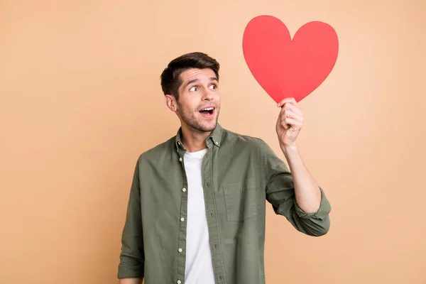 Photo of amazed charming happy man look empty space hold red paper heart isolated on beige color background — Stock Photo, Image
