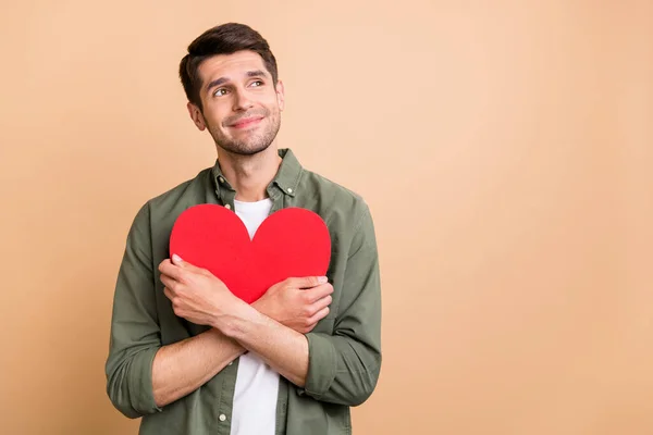 Photo of excited dreamy young guy dressed green shirt embracing red heart looking empty space isolated beige color background — Stock Photo, Image