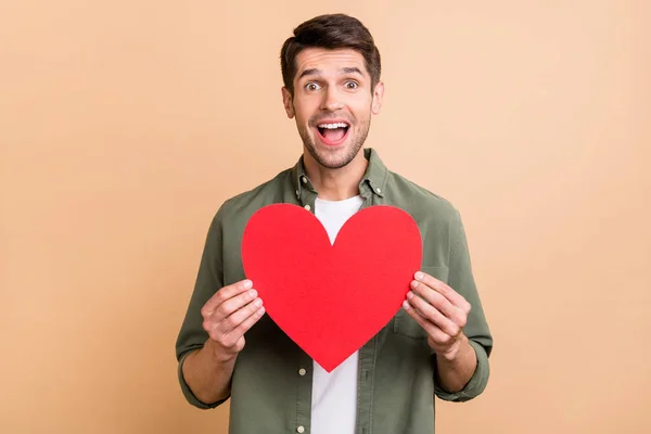 Foto de encantador jovem espantado homem segurar mãos vermelho papel coração forma isolada no fundo cor bege — Fotografia de Stock