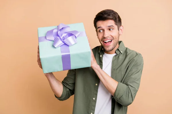 Foto de chico joven emocionado sorprendido vestido camisa verde tratando de adivinar dentro de la caja actual aislado color beige fondo —  Fotos de Stock