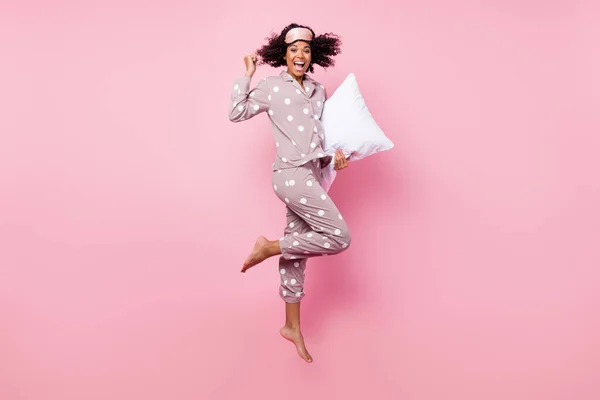 Photo of lucky adorable wavy dark skin lady dotted sleepwear jumping high holding white pillow rising fist isolated pink color background — 스톡 사진