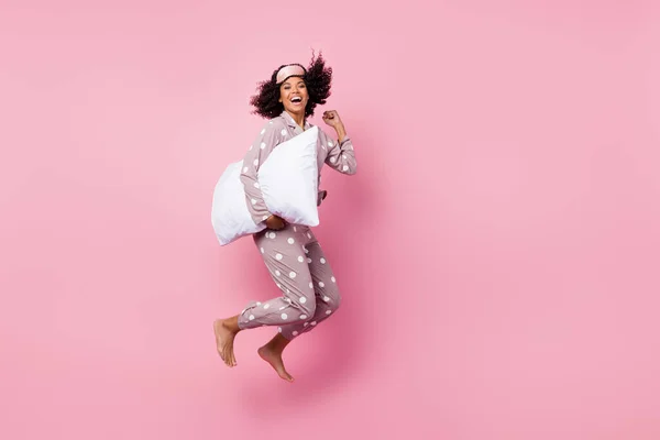 Photo of funky lucky wavy dark skin lady dotted sleepwear jumping high holding white pillow rising fist isolated pink color background — ストック写真