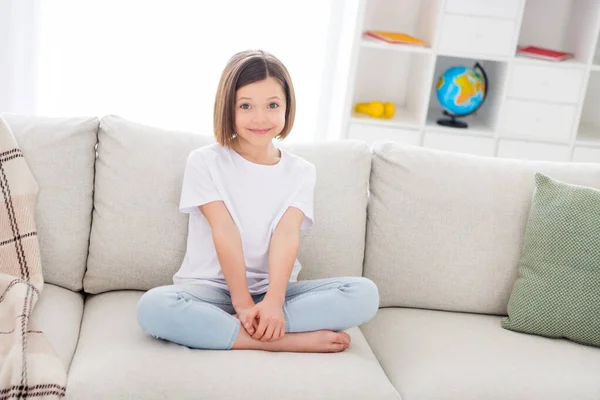 Photo de douce adorable petite écolière habillée blanc t-shirt assis coach jambes croisées à l'intérieur chambre maison maison — Photo