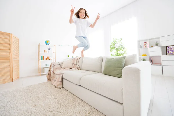 Foto von ziemlich funky kleine Schulmädchen gekleidet weißes T-Shirt zeigt V-Zeichen springen Trainer drinnen Zimmer zu Hause Haus — Stockfoto