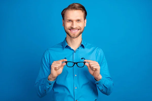 Фото молодого красивого мужчины счастливая положительная улыбка держать очки изолированы на синем фоне цвета — стоковое фото