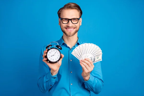 Foto de joven hombre de negocios feliz sonrisa positiva celebrar reloj tiempo dinero en efectivo ricos dólares aislados sobre fondo de color azul —  Fotos de Stock
