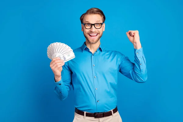 Fotografie mladého vzrušeného muže šťastný pozitivní úsměv držet peníze v hotovosti dolary radovat vítězství loterie pěsti ruku izolované přes modré barvy pozadí — Stock fotografie