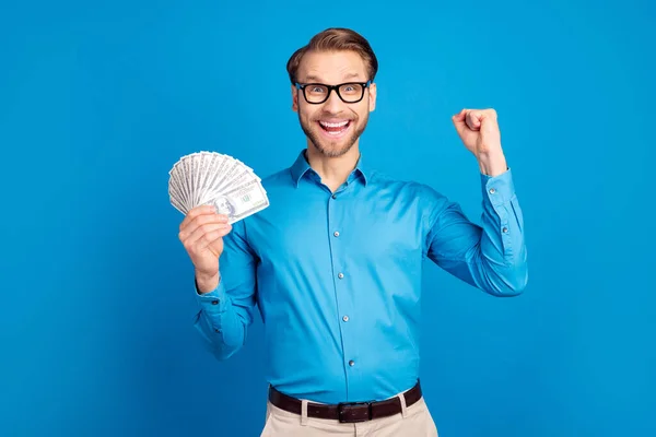 Foto de joven emocionado hombre feliz sonrisa positiva mantener dinero dólares en efectivo regocijarse victoria lotería puños mano aislada sobre fondo de color azul —  Fotos de Stock