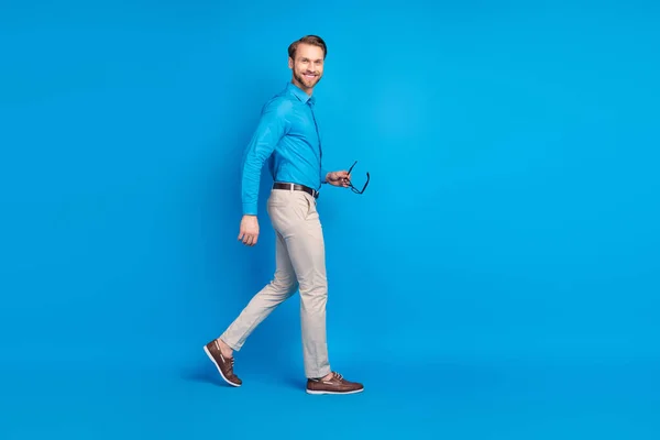 Полный профиль тела боковой фото молодого человека счастливой позитивной улыбкой идти держать очки, изолированные на синем фоне цвета — стоковое фото