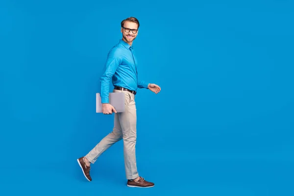 Full length profil boční fotografie mladého obchodníka šťastný pozitivní úsměv jít chodit držet notebook izolované přes modré barvy pozadí — Stock fotografie