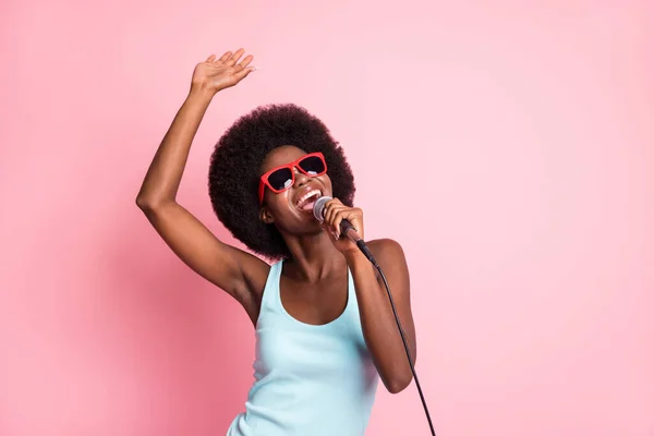 Retrato de optimista bonita morena rizada dama cantar en gafas de desgaste de micrófono top azul aislado sobre fondo de color rosa — Foto de Stock