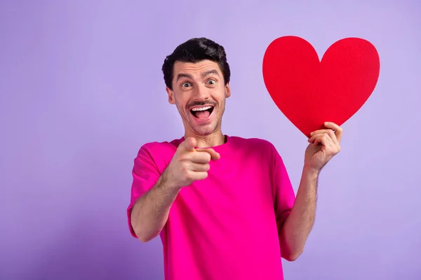 Portrét udiveného chlapa držet papírové srdce pohlednice ukazují prstem fotoaparát jste izolované na fialové barvy pozadí — Stock fotografie