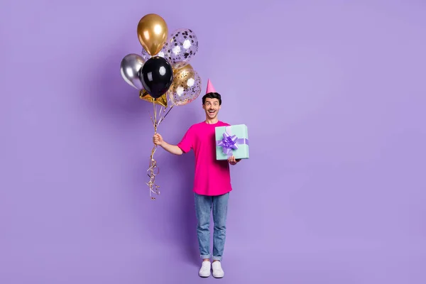 Foto de tamaño completo de joven feliz hombre emocionado mantenga globos usar tapa de cono celebrar regalo de cumpleaños aislado en el fondo de color púrpura —  Fotos de Stock