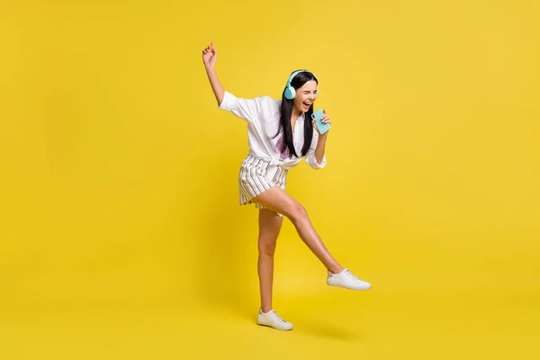 Foto de tamaño completo de la joven alegre sonriente chica positiva en los auriculares cantando en el teléfono aislado en el fondo de color amarillo —  Fotos de Stock