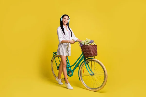 Perfil de corpo inteiro foto lateral da morena na moda mulher segurar bicicleta desgaste fone de ouvido isolado no fundo de cor amarela — Fotografia de Stock