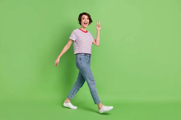 Full längd kroppsstorlek profil sida syn på ganska förvånad glad glad flicka promenader visar v-tecken isolerad över grön pastell färg bakgrund — Stockfoto
