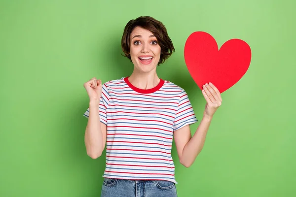 Retrato de menina sortuda alegre atraente segurando na mão cartão de forma de coração se divertindo isolado sobre fundo de cor pastel verde — Fotografia de Stock