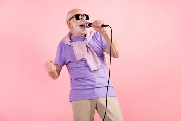 Foto porträtt av funky senior man sjunger på scenen hålla mikrofon bär solglas isolerad på pastell rosa färg bakgrund — Stockfoto