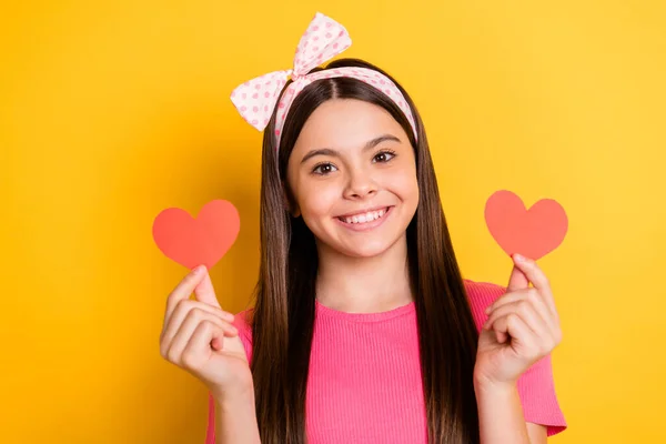 Foto de adorável brilhante pequena estudante vestida camiseta rosa segurando dois corações de papel vermelho sorrindo isolado cor amarela fundo — Fotografia de Stock