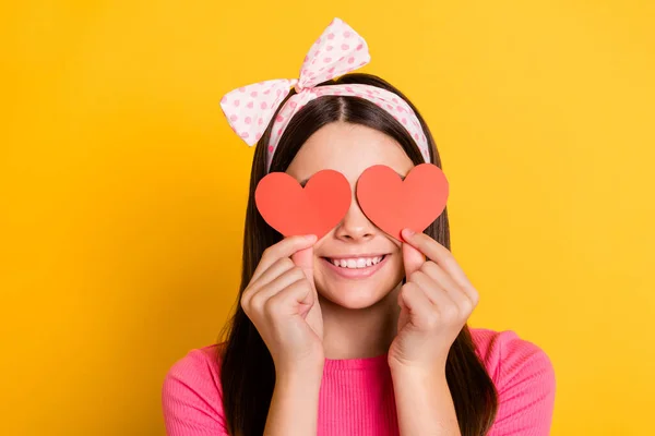 Foto de doce menina bonita escola usar rosa t-shirt sorrindo segurando dois papéis corações vermelhos cobrir os olhos isolado cor amarela fundo — Fotografia de Stock