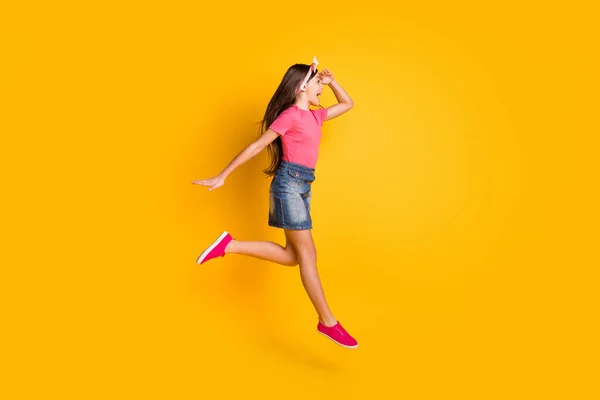 Foto van schattige funky schoolmeisje gekleed roze t-shirt springen hoog lachende arm voorhoofd op zoek lege ruimte geïsoleerde gele kleur achtergrond — Stockfoto