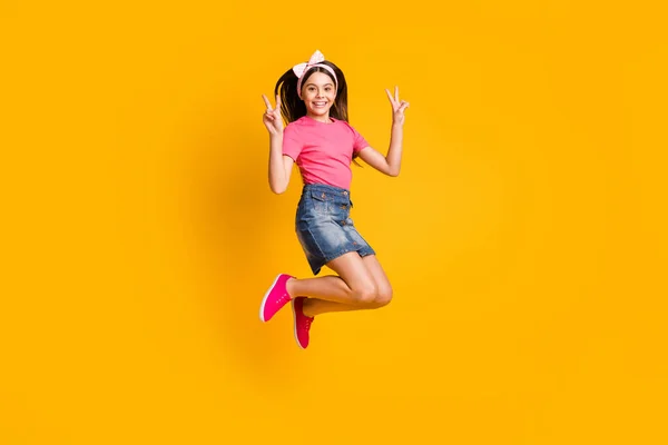 Foto de la pequeña colegiala dulce funky vestida camiseta rosa saltando alto mostrando dos signos de v sonriendo aislado color amarillo de fondo —  Fotos de Stock