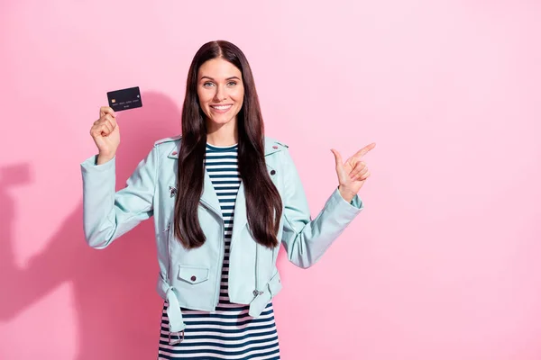 Foto av ung lycklig kvinna bära randig klänning blå läder jacka punkt tomt utrymme hålla kreditkort isolerad på rosa färg bakgrund — Stockfoto