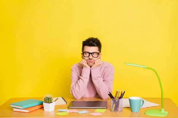 Foto de hombre joven infeliz manos tristes tacto mejillas sentarse mesa aburrido lección aislada sobre fondo de color amarillo — Foto de Stock