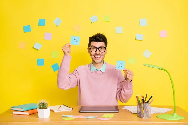 Foto des jungen fröhlich lächelnden positiven Mannes in einer Brille, die glückliche und traurige Papier-Emojis auf gelbem Hintergrund zeigt — Stockfoto