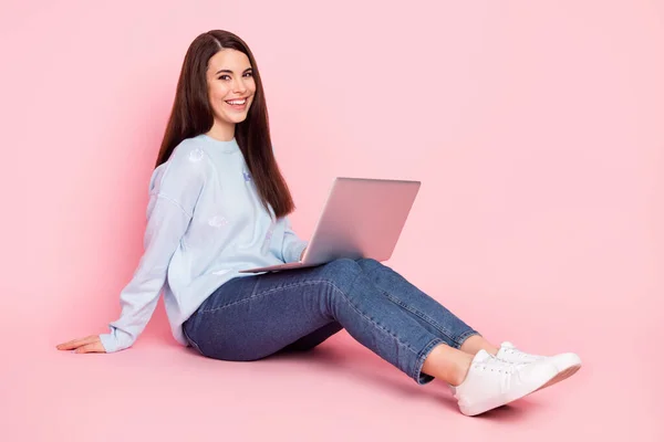 Полноразмерная фотография профиля симпатичной оптимистичной брюнетки с прической, сидящей на ноутбуке в джинсах на розовом фоне — стоковое фото