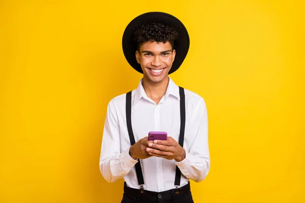 Foto van vrolijke man houden telefoon tand glimlach dragen hoed wit shirt bretels geïsoleerd geel kleur achtergrond — Stockfoto