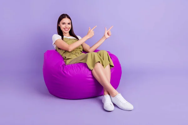 Foto tubuh penuh kebahagiaan wanita muda menunjuk ruang kosong jari duduk kantong kacang terisolasi pada latar belakang warna ungu — Stok Foto