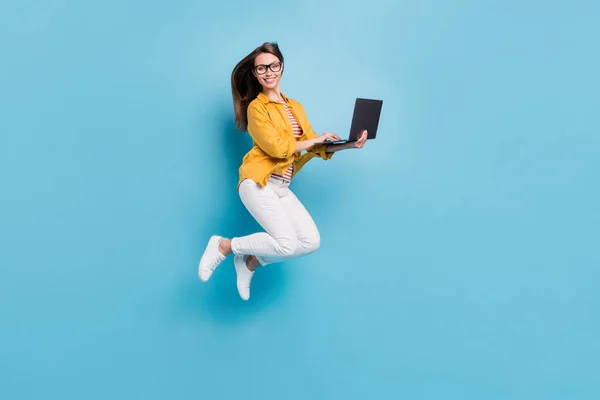 迷人而快乐的年轻女子跳起来的全长照片，手持电脑，用淡蓝色背景隔开 — 图库照片