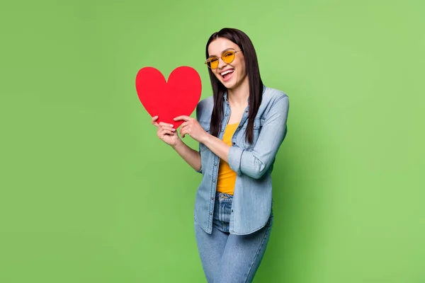 Foto de jovem alegre sorrindo mulher bonita em vidro solar segurar grande coração de papel vermelho isolado no fundo de cor verde — Fotografia de Stock