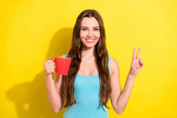 Портрет привлекательной веселой девушки, пьющей горячее какао расслабиться, показывая V-знак изолированы на ярко-желтый цвет фона — стоковое фото