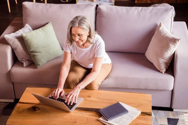 Foto de mulher bonita adorável aposentado vestido branco t-shirt sentado sofá digitando dispositivo moderno sorrindo dentro de casa plana casa — Fotografia de Stock