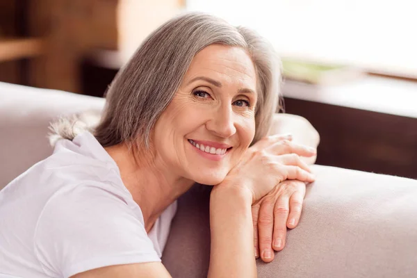 Photo of sweet shiny old woman dressed white t-shirt sitting sofa arms cheeks smiling indoors flat home house — ストック写真