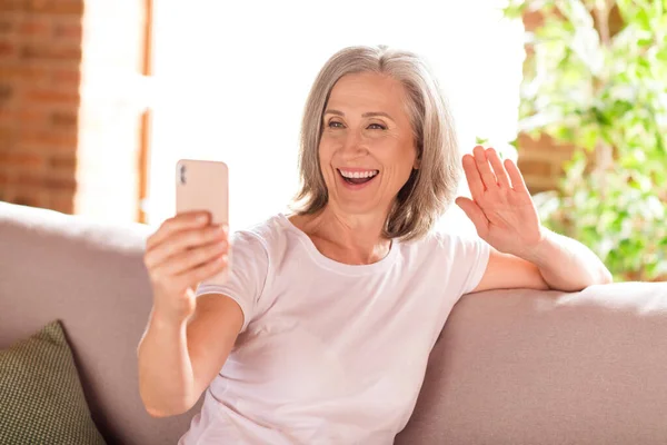 Фото милой хорошенькой пенсионерки носить белые футболки сидя диван улыбаясь говорящий современный гаджет размахивая рукой в помещении дома квартиру — стоковое фото