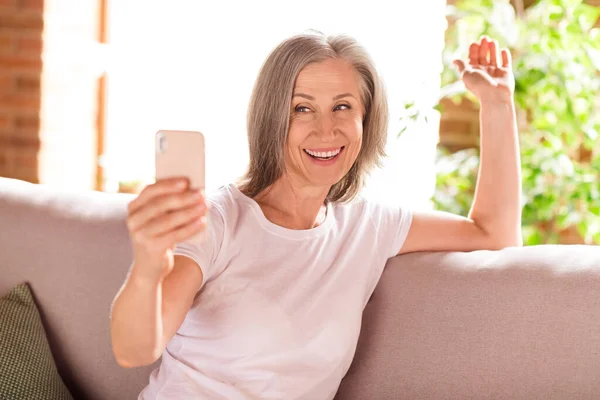 Photo of adorable sweet retired woman dressed white t-shirt sitting couch communicating modern device smiling indoors flat home house — Stok fotoğraf