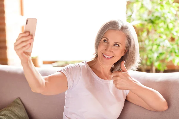 Portrait of elderly retired trendy cheery grey-haired woman sitting on sofa taking selfie showing thumbup at home house indoors — Stok fotoğraf