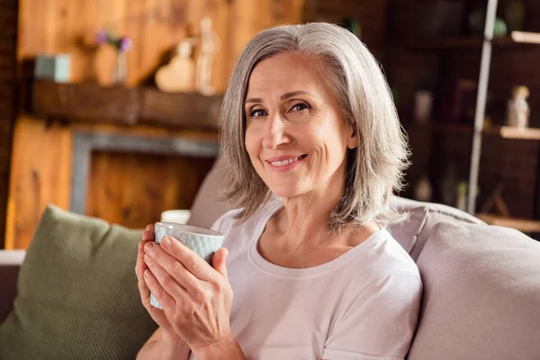 Photo of shiny sweet lady pensioner wear white t-shirt sitting sofa smiling drinking coffee indoors house flat — ストック写真