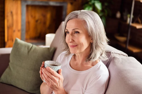 Portrait of attractive dreamy grey-haired woman sitting on divan drinking cacao nice day at home house flat indoors — ストック写真