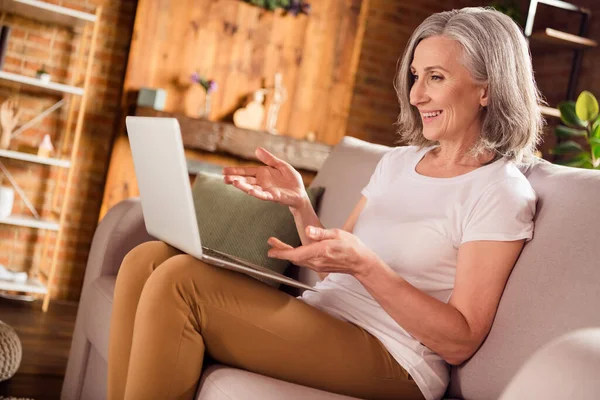 Foto de doce bonita senhora pensioner desgaste branco t-shirt sentado sofá sorrindo falando gadget moderno em casa plana — Fotografia de Stock